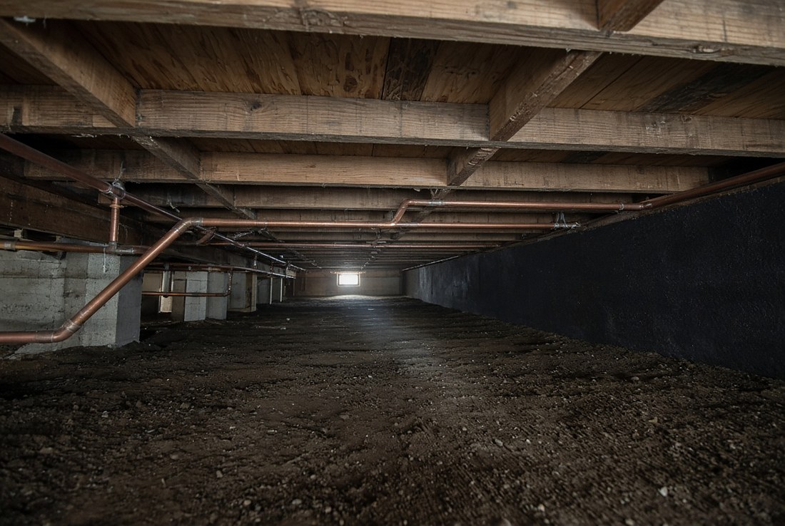 The inside of a vented crawlspace under a house, looking down its length: copper water pipes running beneath wooden floor joists, concrete block piers, a bare dirt floor, and a small foundation vent opening letting daylight in at the far end