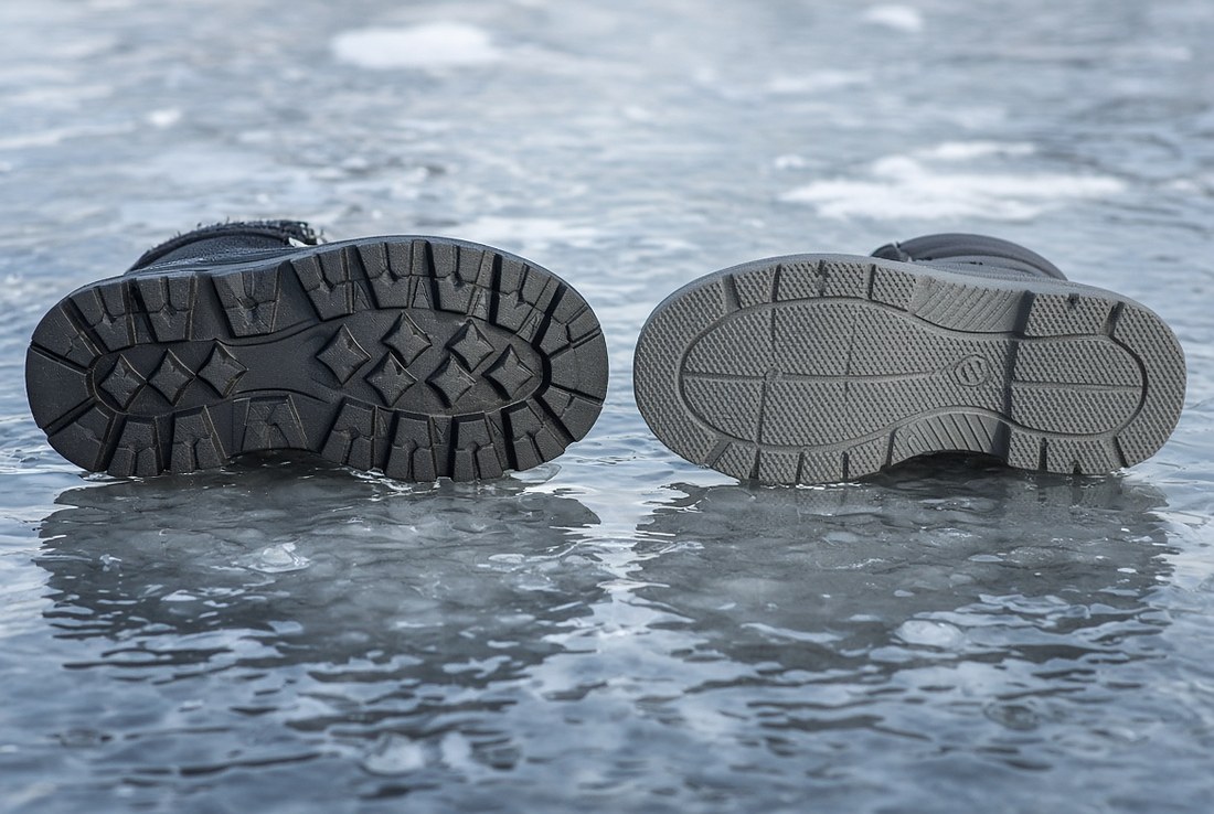 Two winter boots laid on their sides on wet ice with the soles facing the camera, one with deep blocky aggressive lugs and one with a flatter, finer-textured tread pattern