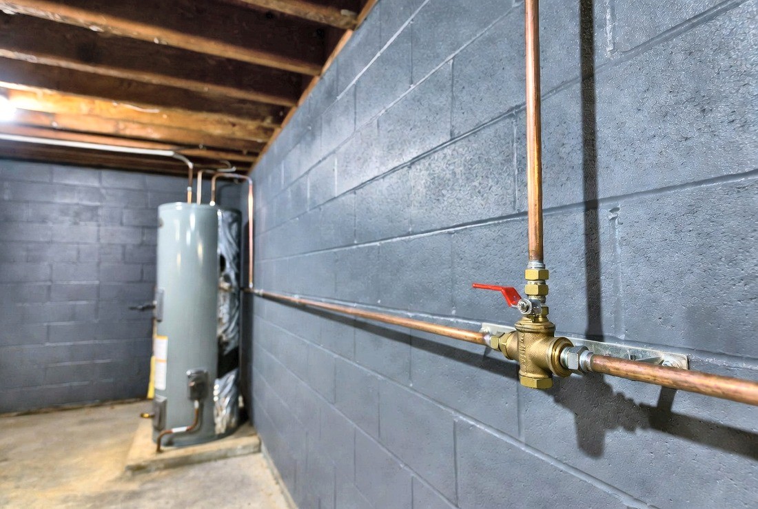 An interior main water shutoff on a basement block wall — copper pipe with a red-handled lever ball valve, the lever turned crosswise to the pipe, a water heater behind it